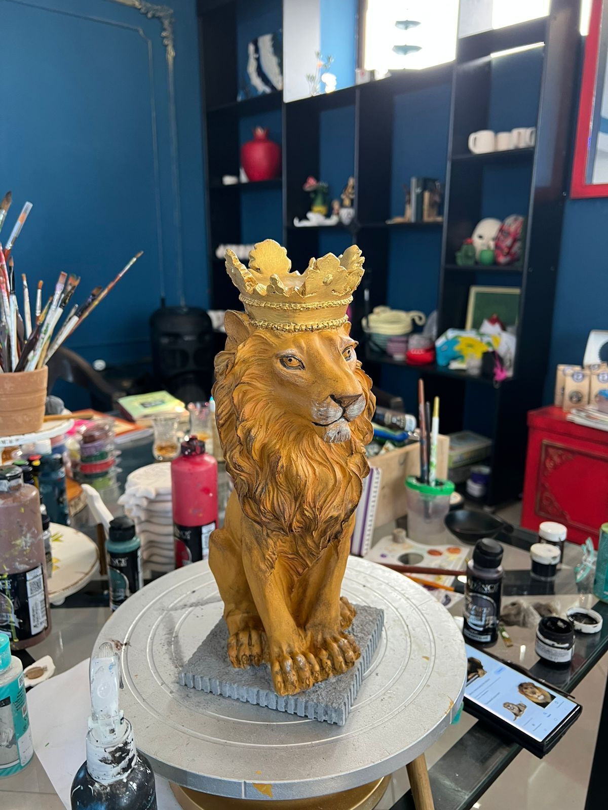 Une sculpture de lion, coiffée d’une couronne, est posée sur un piédestal dans une pièce où se trouvent des étagères et divers objets.