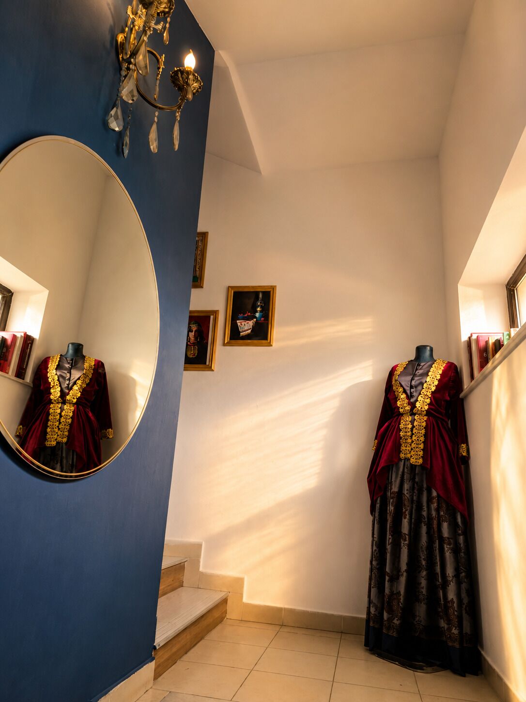 Una habitación con un espejo grande, un maniquí vestido con un atuendo rojo y dorado, un candelabro y obras de arte enmarcadas en la pared.