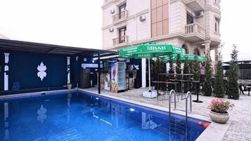 A swimming pool with a building in the background, a covered area with a sign, and outdoor seating.