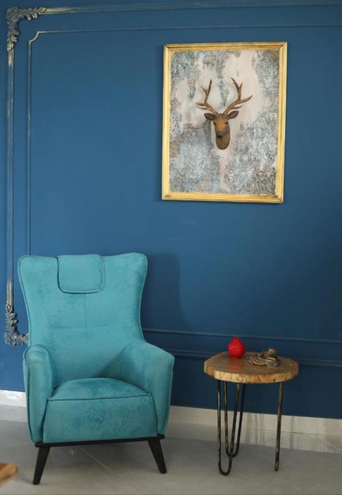A teal armchair, a small wooden table with a red object, and a framed deer head artwork on a blue wall.