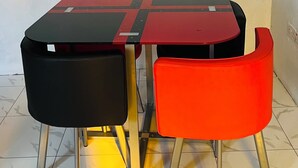 A table with two chairs, one black and one red, in a room with a plain white wall and tiled floor. - OSIM HOMES (Arusha)