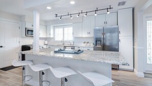 Una cocina moderna con armarios blancos, una isla grande con taburetes de bar, electrodomésticos de acero inoxidable y un ventanal que proporciona luz natural.