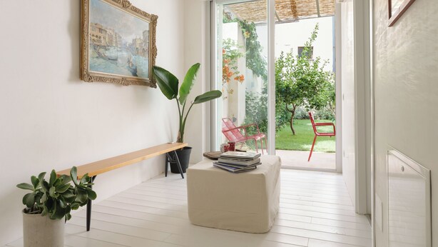 A bright room with a large window, a bench, a potted plant, and a framed painting on the wall.