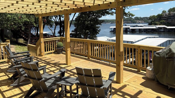 A wooden deck with chairs and a grill, overlooking a lake with docks and boats.
