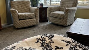 A room with two patterned armchairs, a round ottoman with a floral design, and a view of a body of water through large windows.