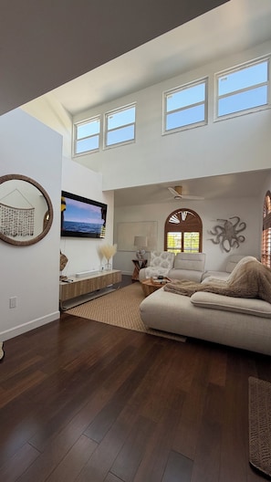 A spacious living room with a large flat-screen TV, a sofa, and a coffee table. There are multiple windows allowing natural light and a ceiling fan.
