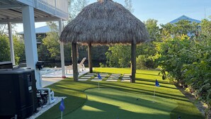 Un césped artificial bien cuidado con un quiosco de paja, una piscina y una zona de estar cubierta.