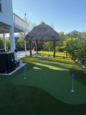 Une pelouse artificielle bien entretenue, agrémentée d’un gazebo à toit de chaume, d’une piscine et d’un espace salon couvert.