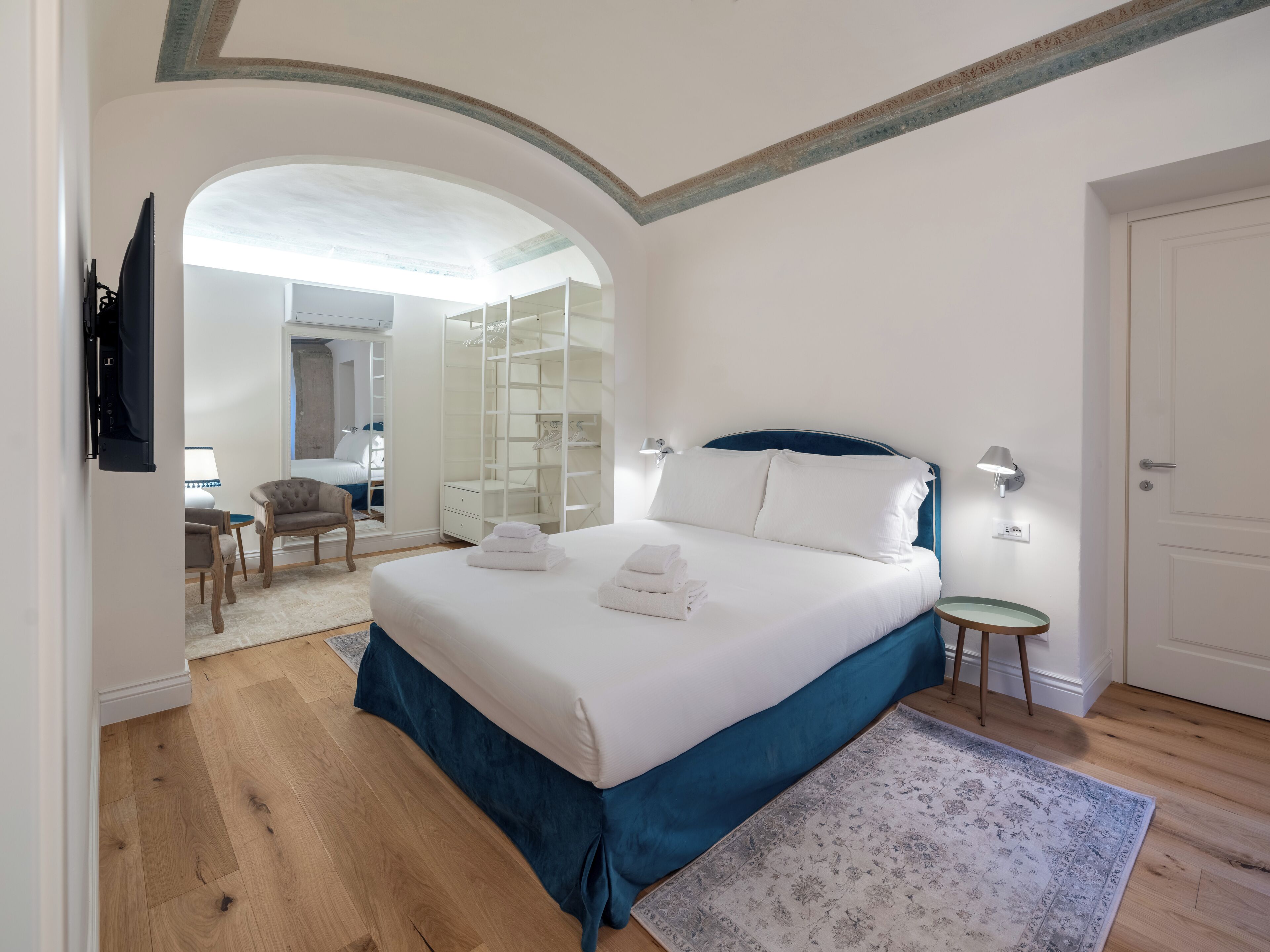 A bedroom with a large bed, a walk-in closet, a desk with a chair, and a view through an arched doorway.