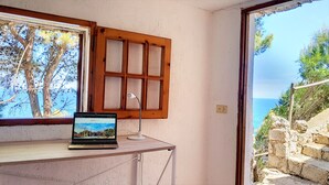 A room with a laptop on a desk, a window with wooden shutters, and a view of the sea and trees. - Charming stone trullo with sea access (house F) (Castro)