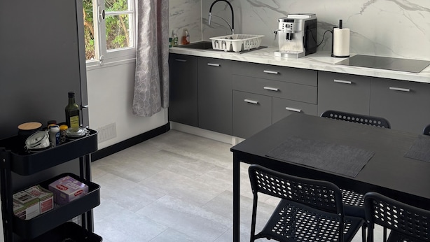 A modern kitchen with grey cabinets, a black countertop, and a built-in oven. There is a window with curtains, a black dining table with chairs, and a black cart with items on it. - Quiet house with garden (Alès)