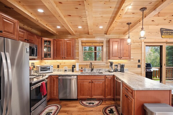 A wooden cabin kitchen with stainless steel appliances, wooden cabinets, and a large window. - Riverfront, Brand New Log Cabin-Fireplace-Firepit (Carroll)