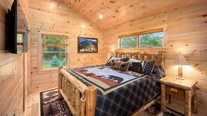 Un dormitorio de cabaña de madera acogedor, con cama, dos ventanas, un televisor montado y un cuadro en la pared.