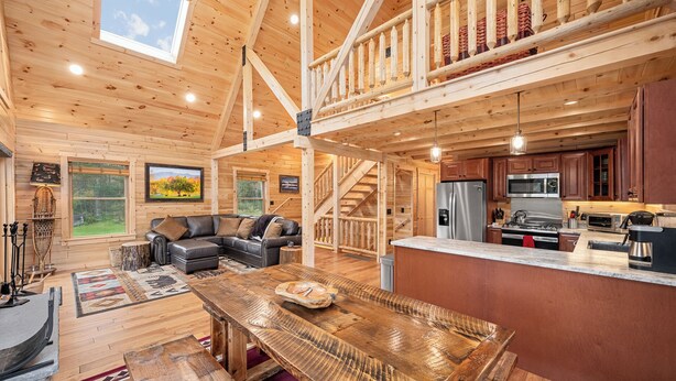 Un interior de cabaña espacioso con detalles de madera, un comedor, una cocina y una sala de estar con un ventanal que da a un bosque.