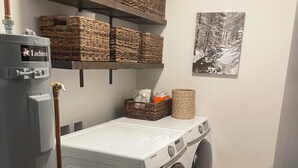 A laundry room with a front-loading washer and dryer, a water heater, and wicker baskets on shelves. - Your Bozeman Basecamp: Comfortable Condo in the Perfect Downtown Location (Bozeman)
