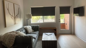 A modern living room with a grey sofa, a coffee table, and a large window with blinds. - Your Bozeman Basecamp: Comfortable Condo in the Perfect Downtown Location (Bozeman)
