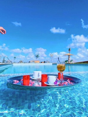 A poolside setting with a tray of drinks and cups, palm trees, and a clear blue sky.