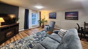 A living room with a sofa, a TV, a lamp, a plant, and a view of the outdoors. - Spacious Apartment in Horseheads, NY (Horseheads)