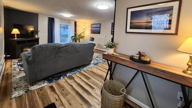 A modern living room with a grey sofa, wooden flooring, a blue rug, a wooden side table with a lamp, and a framed picture on the wall. - Spacious Apartment in Horseheads, NY (Horseheads)