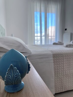 Un dormitorio con cama, ropa de cama blanca, una ventana con cortinas y un objeto decorativo azul sobre una superficie de madera.