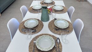 A dining area with a long table set for six, white chairs, and a TV in the background.