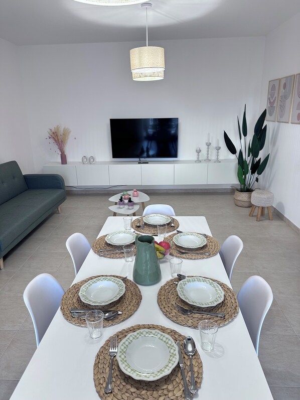 Un comedor con una mesa larga dispuesta para seis personas, sillas blancas y un televisor al fondo.
