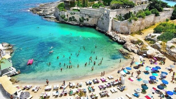 A coastal area with a beach, clear turquoise waters, and a fortified structure with greenery.