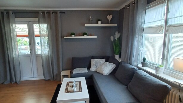En moderne stue med en grå sofa, et hvidt sofabord og en sort måtte. Der er hvide reoler med dekorative genstande, og et vindue med gardiner.