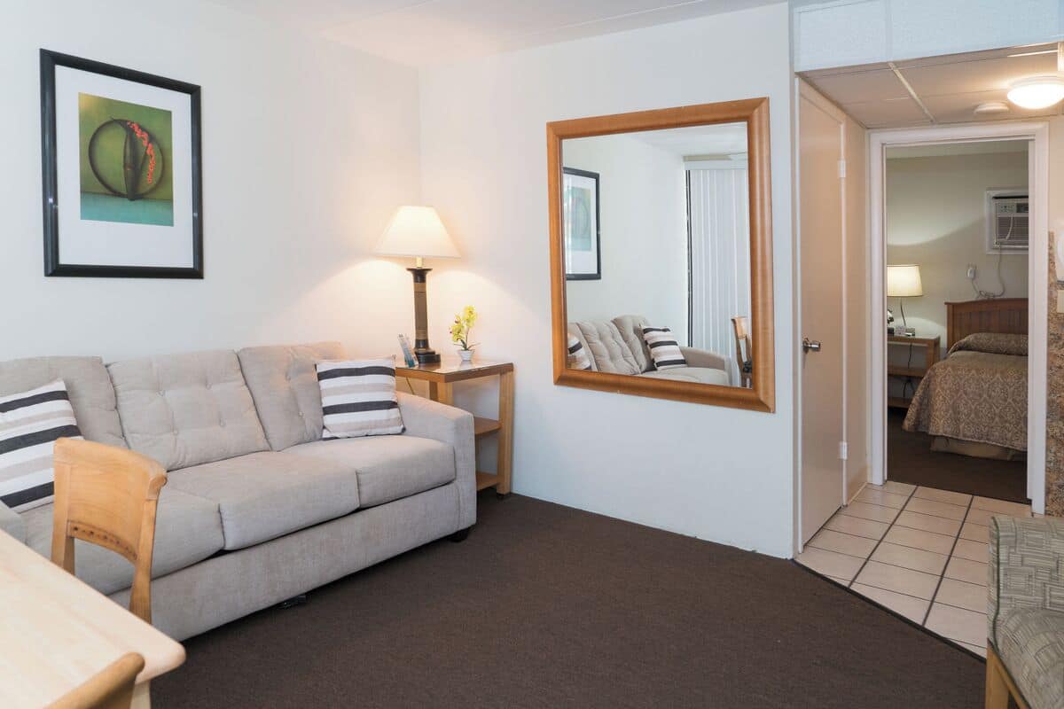 A living room with a sofa, a table, a lamp, a mirror, and a door leading to a bedroom.