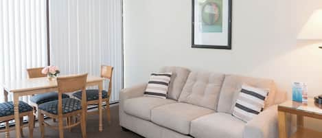 Una sala de estar con un sofá color beige, almohadones a rayas, una mesa de comedor de madera con sillas, un ventilador de techo y un cuadro en la pared.