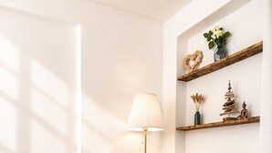Ein Zimmer mit zwei beigen Sesseln, einer Stehlampe, einem kleinen Tisch mit einer grünen Schale und einem Wandschrank mit Dekorationen.