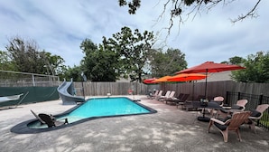 Un jardin avec une piscine, des chaises longues et une table ombragée par des parasols.