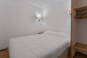 A bedroom with a single bed, a nightstand, a wall-mounted lamp, and a wooden wardrobe with hanging clothes.