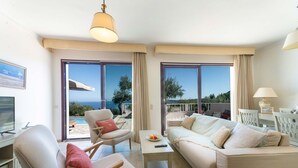 Ein geräumiges Wohnzimmer mit einer Couch, Sesseln, einem Couchtisch und einem Fernseher. Große Fenster bieten einen Ausblick ins Freie.