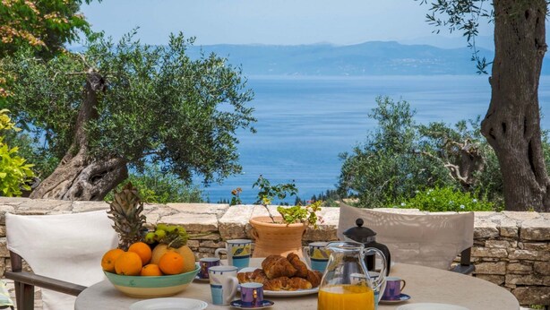 A table set with breakfast items, including fruits, pastries, and a jug of orange juice, overlooking a scenic view of the sea. - Amara Villa - Three Bedroom Villa, Sleeps 6 (Paxi)