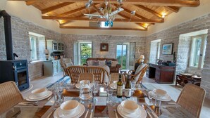 A dining area with a glass table set for six, surrounded by wicker chairs, with a stone wall and wooden ceiling beams in the background. - Amara Villa - Three Bedroom Villa, Sleeps 6 (Paxi)