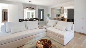 En moderne stue med en hvid hjørnesofa, et sofabord i glas med dekorative genstande, og et køkken i baggrunden.