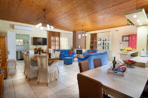 Un vaste espace de vie doté d’un plafond en bois, d’une table à manger avec des chaises, de canapés bleus et d’un coin cuisine avec une télévision et un panier de fruits.