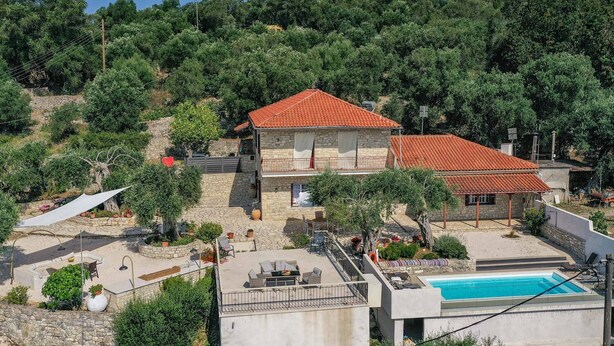 A two-story stone house with a red-tiled roof, a swimming pool, and a patio area with outdoor furniture.