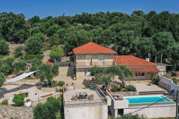 A two-story stone house with a red-tiled roof, a swimming pool, and a patio area with outdoor furniture.