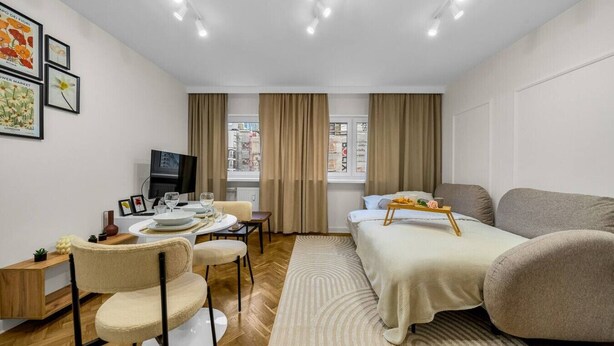 A modern hotel room with a bed, a sofa, a dining area with a table and chairs, a TV, and a window with curtains.