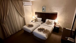 Ett hotellrum med två sängar, ett stort fönster med gardiner, en luftkonditioneringsanläggning, ett nattduksbord med en lampa och en telefon.
