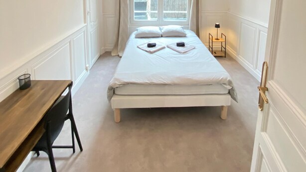 A bedroom with a bed, a desk, a lamp, and a window with curtains.