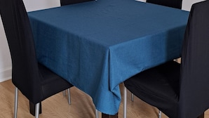 Una habitación con una mesa cubierta por un mantel azul, cuatro sillas negras y piso de madera.