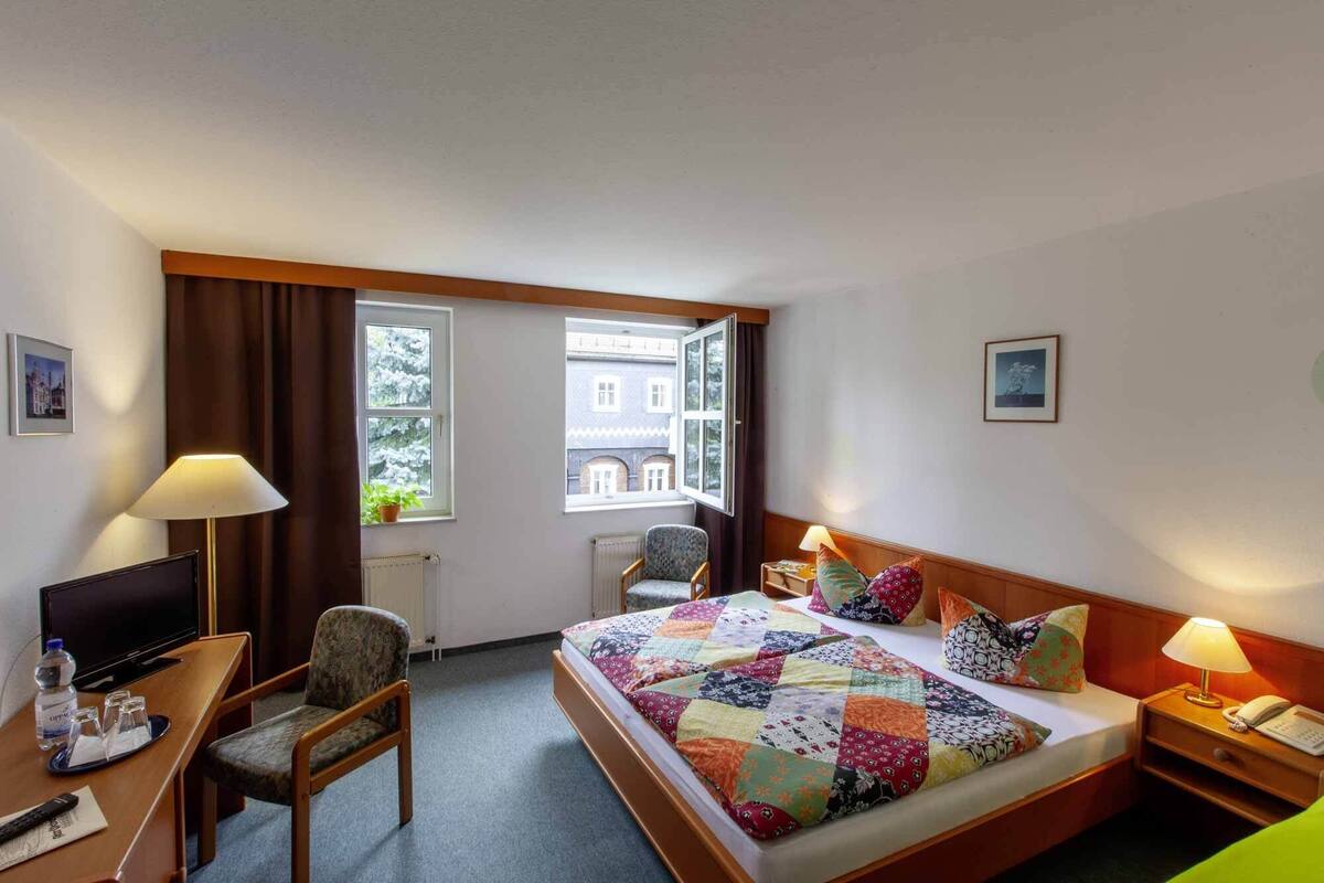 Habitación de hotel con cama, escritorio, silla, televisión y dos ventanas.