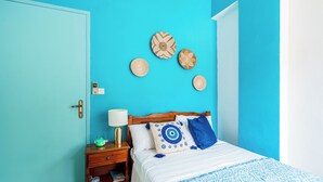 A room with a turquoise wall, a wooden bed with a blue and white patterned bedspread, a nightstand with a lamp, and three woven wall decorations. - Double Room Apartment (Poros kefalonias)