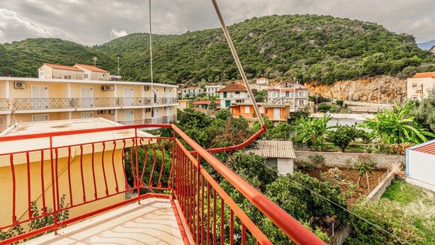 A coastal residential area with houses, a balcony with red railings, and a mountainous backdrop. - Double Room Apartment (Poros kefalonias)