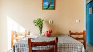 A dining area with a checkered tablecloth, wooden chairs, a vase with green foliage, and a bowl of fruit. - Double Room Apartment (Poros kefalonias)