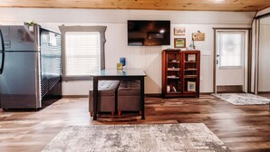 Amplia sala de estar con techo de madera, un televisor de pantalla plana grande montado en la pared, un refrigerador moderno, un mueble de madera con libros y una mesa de comedor con sillas.