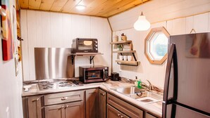 Cocina compacta con estufa, microondas, tostadora y refrigerador. Hay repisas de madera y una ventana sobre el fregadero.